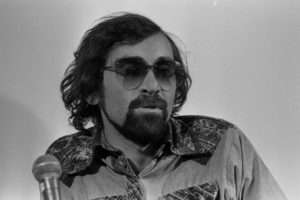 Man with dark curly hair and beard wearing aviator sunglasses and print shirt behind silver microphone